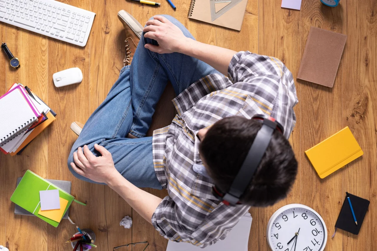 پسربچه نشسته روی زمین چوبی با هدفون روی سر و دفترچه ها و ساعت روی زمین